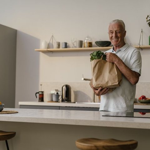 Older man smiling in a kitchen holding a paper bag of groceries